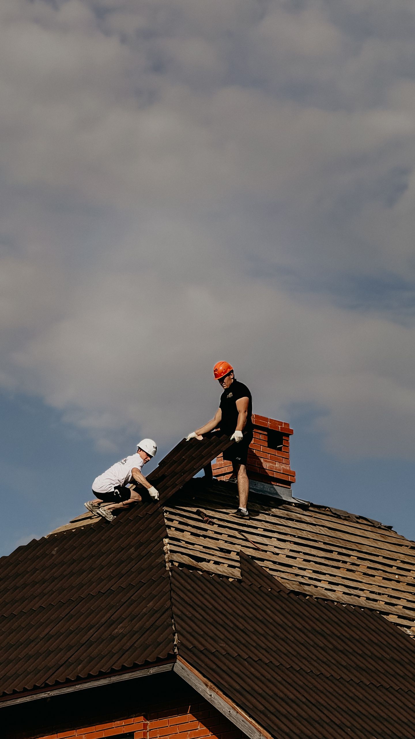 Two workers in safety helmets repair a roof, showcasing outdoor construction during a cloudy day.